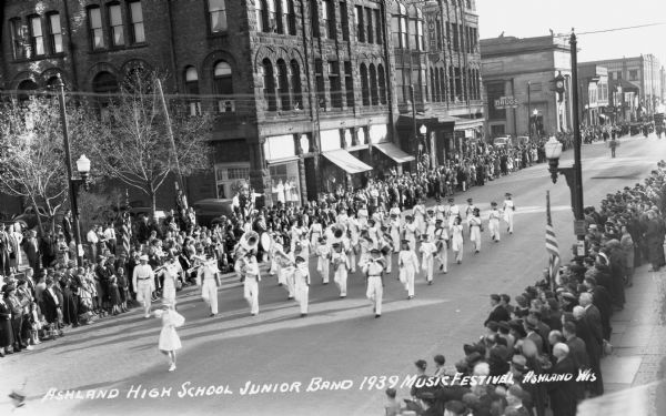 Elevated view of students from the Ashland High School Junior Band at the Music Festival, marching down Main Street. Students are wearing light uniforms or white clothing and are playing instruments including trombones, drums, tubas, and clarinets. Crowds of people are on the sidewalks, and the Knight Hotel is across the street.