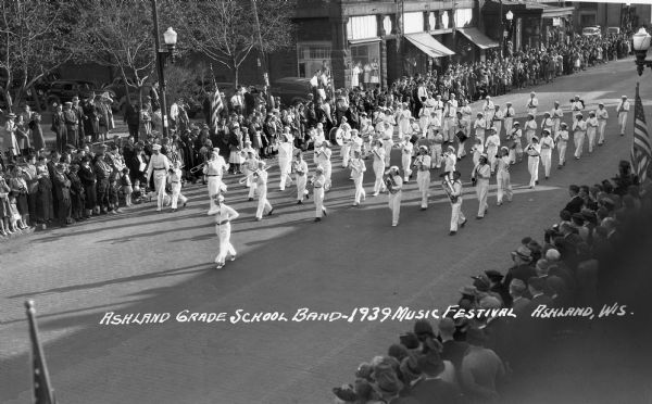 Elevated view of students from the Ashland Grade School Rhythm Band at the Music Festival, marching down Main Street. The students are wearing light uniforms or white clothing and are playing instruments including trombones, drums, trumpets, saxophones, euphoniums, and clarinets.  Crowds of people line the street, and the Knight Hotel is across the street.
