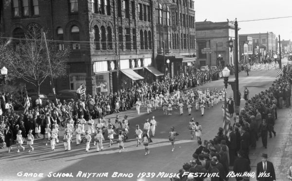 Elevated view of students from the Grade School Rhythm Band at the Music Festival, marching down Main Street. Students are wearing light or white clothing and are playing drums and percussion instruments.  Crowds of people line the street, and the sign on the brownstone building across the street says “Knight Hotel.”