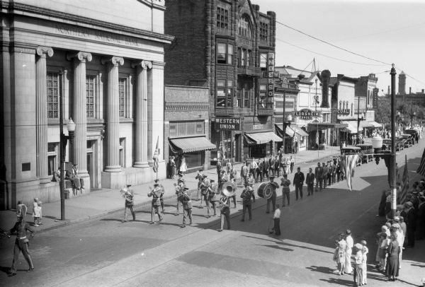 Elevated view of the Bodin Finstad Marching Band on West Main Street. Sixteen men wearing marching band uniforms are playing instruments including euphoniums, a tuba, trombones, a bass drum, trumpets, clarinets, and French horns. The bass drum says "Bodin Finstad Bayfield County, Post 86, Washburn, Wisc." People are standing on the sidewalk watching the parade. Behind the marching band are more men and automobiles. Buildings on the other side of the street include Northern National Bank, Western Union, Lew Anderson, Farm Lands Insurance, Ashland Shoe Store, The Florsheim Shoe, The Majestic, Coal, and JC Penney Co. (JC Penney Co. moved from this location by 1937.)