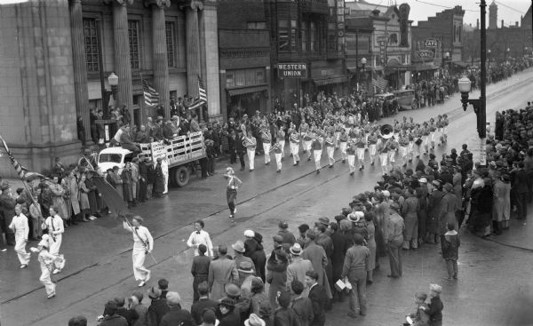 Elevated view of students in a school marching band in a parade marching down Main Street. Students are wearing uniforms and are playing instruments including trombones, drums, tubas, French horn, saxophones, trumpets, and clarinets. Crowds of people line the street, and signs on the buildings in the background on the left read “Western Union,” Schillers Shoes,” “Coal,” and “Marigold Café.”  There is a truck on the far left that says "Central Lumber and Supply Co." that has people standing in the back and has two American flags.