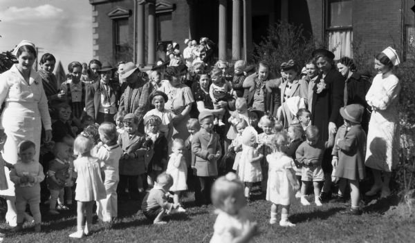 Crowd of women, babies, boys, girls, and nurses standing in front of the Hospital, which was also known as the Wilmarth Mansion. Some of the women are wearing hats and coats and some are wearing white nurse’s uniforms.