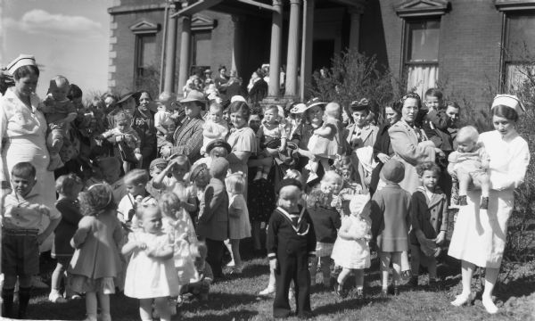 Crowd of women, babies, boys, girls, and nurses standing in front of the Hospital, which was also known as the Wilmarth Masion. Some of the women are wearing hats and coats and some are wearing white nurses uniforms.