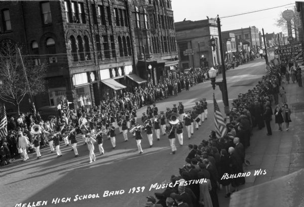 Elevated view of the Mellen High School Marching Band in the Music Festival parade marching down Main Street. Students are wearing marching band uniforms and are playing instruments including trombones, drums, tubas, trumpets, and clarinets. Crowds of people line the street, and signs on the buildings in the background on the left read "Dr. Forster Dentist," "Knight Hotel," and "Knight Block, Drugs, Cigars, Candy, Soda." Signs on the buildings on the right show “Ashland Cleaners, Dyers” and “The Coffee Pot, Booths.”