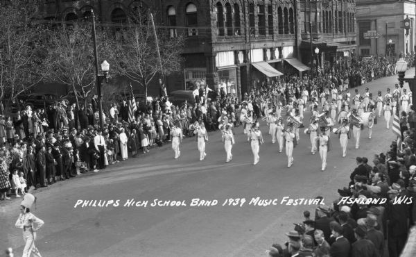 Elevated view of the Phillips High School Marching Band in the Music Festival parade marching down Main Street. Students are wearing marching band uniforms and are playing instruments including trombones, drums, tubas, french horns and clarinets. Crowds of people line the street, and signs on the buildings in the background read "Dr. Forster Dentist," "Knight Hotel," and "Knight Block, Drugs, Cigars, Candy, Soda."