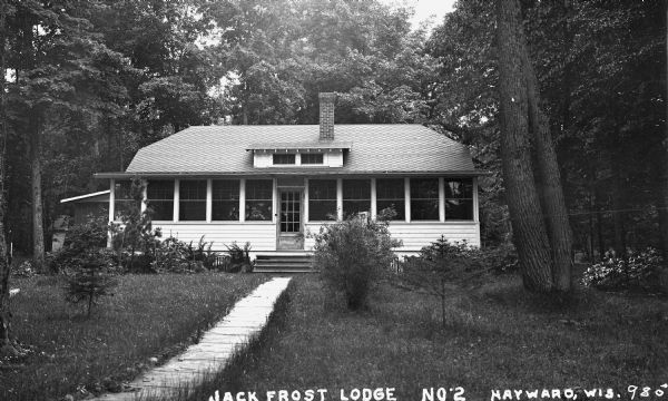 Exterior view of the front facade showing the main doorway and front porch on the Jack Frost Lodge No. 2 on Round Lake. The building has one brick chimney and a sidewalk or path that leads up to the front door.