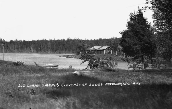 View across field of a one-story log cabin, Smead's Cloverleaf Lodge, across Teal Lake. There is a wooden dock across the lake in front of the lodge and a dock in the foreground with two boats.