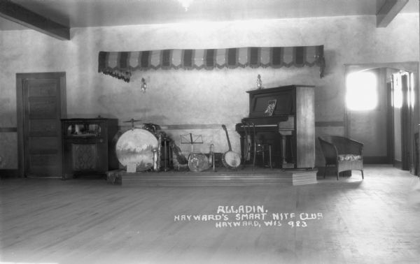 Interior view of Alladin, Hayward's Smart Nite Club showing musical instruments on the stage. Instruments include, from left to right, a drum set, saxophone, fiddle, possibly a guitar or bass, trumpet, banjo, and upright piano.
