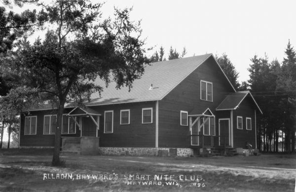 Exterior view of the one-story wooden building with a stone foundation called Alladin, Hayward's Smart Nite Club, with pine trees in the background.