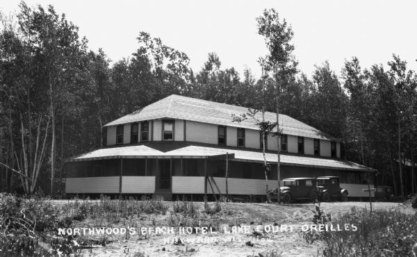 Two automobiles parked outside the two-story wooden building, which has a wrap around screened in porch on the first floor. This building is the Northwood's Beach Hotel on Lake Court Oreilles.