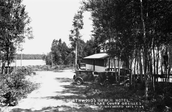 View down drive towards lake. The front porch is behind a grove of trees where automobiles are parked out front of the Northwood's Beach Hotel on Lake Court Oreilles.