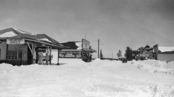 View looking down a very snowy Main Street. Main Street businesses visible on the left include Tire and Battery Service Marathon Tires. A group of people is gathered in front of Hunt’s Cash Store “Where Cleanliness Predominates.” Other one-story buildings are visible on the right and an automobile is parked on the road.