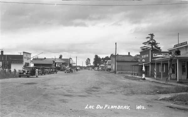 View looking down middle of Main Street. Businesses from left to right include the U.S. Post Office, Tire & Battery Service, Store - Tourist & Supplies, Standard Service, Meats, Gift Shoppe, Marshfield Beer, … Drop Inn, and The Gauthier Store. There are 13 automobiles along Main Street. Two men are working on the roof of the Gauthier’s Store on the far right.