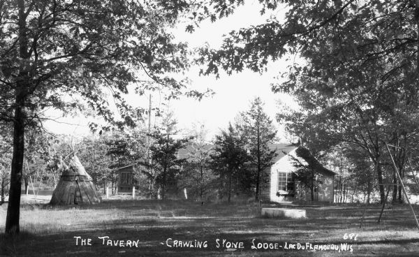 View of a one-story log lodge, the Tavern - Crawling Stone Lodge, and a tipi with trees in the background and foreground. There is a lake behind the lodge.