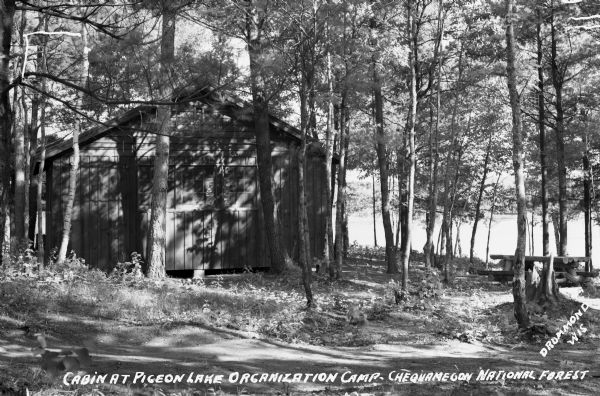 View of a cabin and picnic table at Pigeon Lake Organization Camp in the Chequamegon National Forest. The cabin is surrounded by trees, and in the distance the lake is visible.