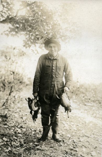 Outdoor portrait of a man wearing a hat, jumpsuit, and mud boots holding a gun and ducks. He is identified as the owner of Nature Lovers Paradise resort on Jackson Lake. The text describing this photograph says, "The owner and writer of this letter last fall."<p>Located within the Chequamegon National Forest, Jackson Lake is a 142-acre drainage lake with a navigable outlet channel to Namakagon Lake – which is a 3,227 acre drainage lake at the headwaters of the Namakagon River in Bayfield County, Wisconsin.</p>