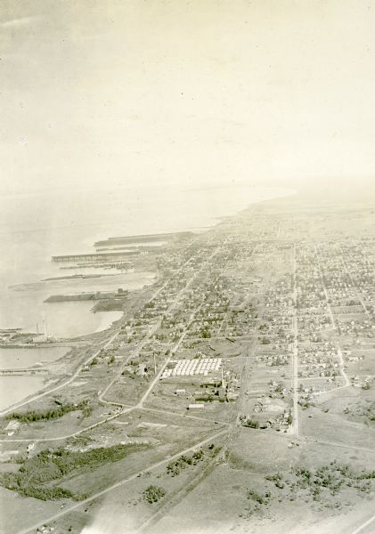 Aerial view of town, showing the shoreline of Lake Superior, ore docks, roads, houses, buildings, trees, and smokestacks. Text describing this photograph says, "City of Ashland; our nearest town of any consequence."