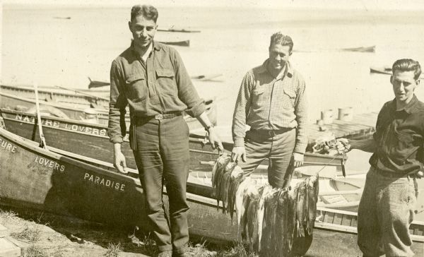 View of three men wearing pants, belts and long sleeve button up shirts holding a stringer of fish they caught. The wooden boats behind them say Nature Lovers Paradise. Behind the men is a dock on Jackson Lake.  Text describing this photograph says, "fish caught here."<p>Located within the Chequamegon National Forest, Jackson Lake is a 142-acre drainage lake with a navigable outlet channel to Namakagon Lake – which is a 3,227 acre drainage lake at the headwaters of the Namakagon River in Bayfield County, Wisconsin.</p>