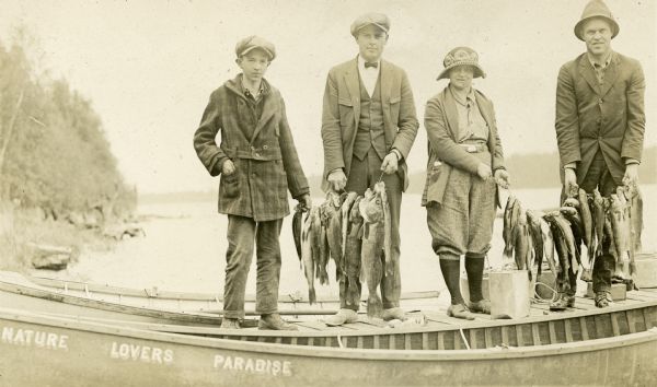Group portrait of four people standing on a pier holding two lines of fish that they caught at Nature Lovers Paradise resort on Jackson Lake. The two men, one woman, and one boy are all wearing hats and either sweaters or coats. The wooden canoe in the foreground says: "Nature Lovers Paradise."<p>Located within the Chequamegon National Forest, Jackson Lake is a 142-acre drainage lake with a navigable outlet channel to Namakagon Lake – which is a 3,227 acre drainage lake at the headwaters of the Namakagon River in Bayfield County, Wisconsin.</p>