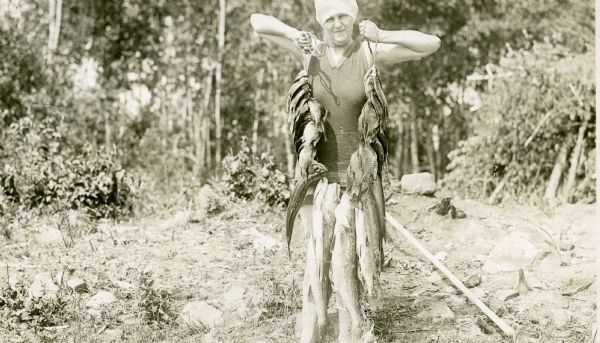 View of a woman wearing a bathing suit and hat holding up a heavy line full of fish caught at Nature Lovers Paradise resort on Jackson Lake.<p>Located within the Chequamegon National Forest, Jackson Lake is a 142-acre drainage lake with a navigable outlet channel to Namakagon Lake – which is a 3,227 acre drainage lake at the headwaters of the Namakagon River in Bayfield County, Wisconsin.</p>