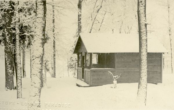 Exterior view of a small one-story wooden cottage or cabin with a front porch, covered in a layer of snow and surrounded by trees on land owned by Nature Lovers Paradise resort on Jackson Lake.<p>Located within the Chequamegon National Forest, Jackson Lake is a 142-acre drainage lake with a navigable outlet channel to Namakagon Lake – which is a 3,227 acre drainage lake at the headwaters of the Namakagon River in Bayfield County, Wisconsin.</p>