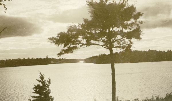 Elevated view of the sun shining through clouds over a lake, identified as Lake Namakagon. In the foreground are trees, and more trees are visible on the opposite shorelines. Text describing the photograph says, "the scenery photos are scenes on Lake Namakagon, all can be reached by rowboat from our cottages."