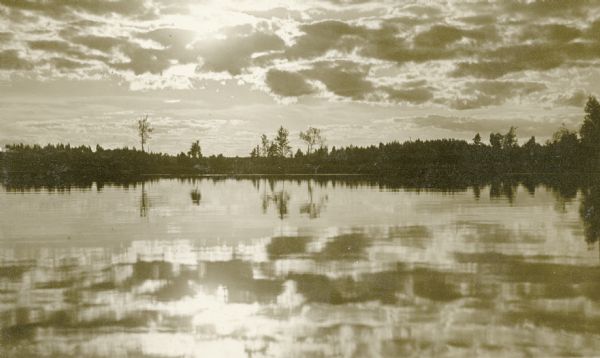 View of the sun shining through clouds over Lake Namakagon. There are trees are visible on the opposite shorelines. Text describing the photograph says, "the scenery photos are scenes on Lake Namakagon, all can be reached by rowboat from our cottages."