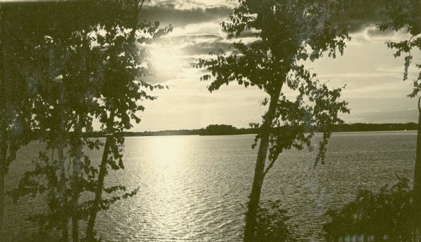 Elevated view of the sun shining through clouds over Lake Namakagon. In the foreground are a few trees and in the distance is the opposite shoreline. Text describing the photograph says, "the scenery photos are scenes on Lake Namakagon, all can be reached by rowboat from our cottages."