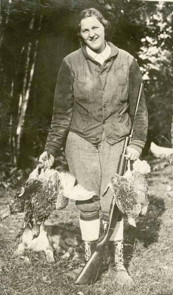 View of a woman standing outdoors holding grouse and a gun. She is wearing boots and a jacket. Behind her is a dog sitting in the grass and trees. Text describing the photograph reads: "Some grouse shot in hunting season."