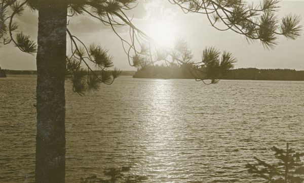 Scenic view of the sun through clouds over a lake, identified as Lake Namakagon. In the foreground is a pine tree, and across the lake the opposite shoreline is visible. Text describes this photograph as, "scenes on Lake Namakagon, all can be reached by rowboat from our cottages" at Nature Lovers Paradise.