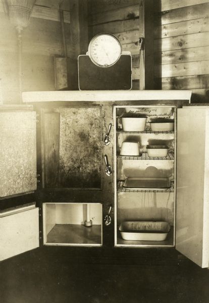 Interior view showing an ice box in a cottage at Nature Lovers Paradise, a resort on Jackson Lake. The doors are open on the ice box, and trays are on the shelves inside. On top of the ice box is a scale. Text below the photographs says, "An icebox like this in each cottage; also white enamel food trays in each ice box.  healthometer scale in each cottage so guests can see from day to day whether they are gaining or losing."