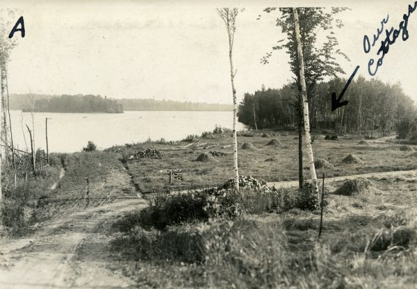 View of a dirt road, cleared land, Jackson Lake and shoreline. Off to the far right is where the Nature Lovers Paradise resort cottages were located. A sign near the road in the shape of an arrow says: Nature Lovers Paradise." Text describing this photograph says, "shows location of cottages; also the beautiful island right out in front."