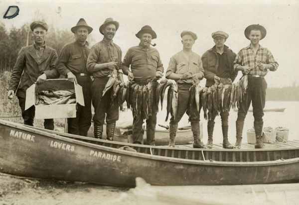 View of seven men wearing pants, boots, belts and long-sleeved button-up shirts holding a box and three stringers of fish they caught. The wooden boats in front of them say "Nature Lovers Paradise". The men are standing on a dock, and in the background is a wooden row boat, and Jackson Lake. Text describing this photograph says, "a morning catch of fish."