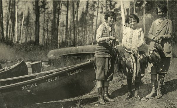 Group portrait of three young women wearing boots standing outdoors and holding onto a heavy line of fish. On the left are some wooden boats, the one in the foreground says "Nature Lovers Paradise." The fish were caught at Nature Lovers Paradise resort located on Jackson Lake.