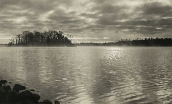 View of the sun shining through clouds over a lake, identified as Lake Namakagon. In the foreground are some rocks on the shoreline, and more trees are visible on the opposite shorelines. There is a fog or mist over the lake. Text describing the photograph says, "the scenery photos are scenes on Lake Namakagon, all can be reached by rowboat from our cottages."