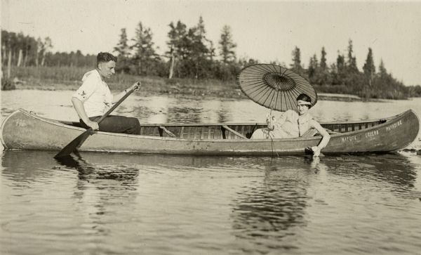 View of a man paddling a canoe with a woman sitting in the front holding an umbrella and white water lily. The canoe says "Nature Lovers Paradise." Text describing this photograph says, "white water lilies grow on our land." Nature Lovers Paradise resort was located on Jackson Lake near Grand View, Wisconsin.<p>Located within the Chequamegon National Forest, Jackson Lake is a 142-acre drainage lake with a navigable outlet channel to Namakagon Lake – which is a 3,227 acre drainage lake at the headwaters of the Namakagon River in Bayfield County, Wisconsin.</p>