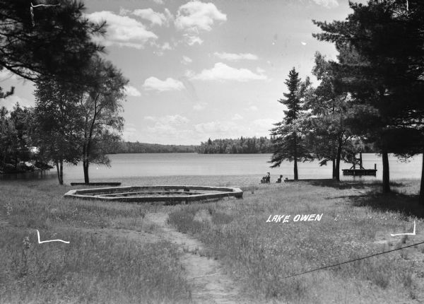 View of a wooden octagonal ring on the shore of Lake Owen. In the foreground are trees and grass. On the right are people on the shore of Lake Owen, and a swimming or diving platform is just offshore. In the distance the opposite shoreline of Lake Owen is visible.