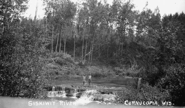 View of two children standing in the Siskiwit River at the top of Bridge Falls. A woman stands on the shore on the right. There are trees and brush on both sides of the river.