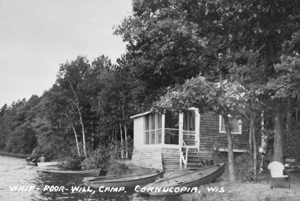 Cedar shake and log cabin near lakeshore with boats. There is a man in a motorboat along the shore.