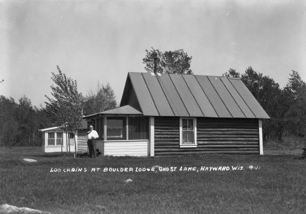Man smoking a cigar while standing in front of a log cabin situated in a clearing in northern Wisconsin. The man's necktie is blowing in the wind.