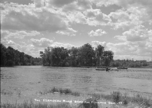 View from shoreline of men fishing out of wooden rowboats on the Flambeau River below the flowage dam. One man is standing in the river. A shack is visible on the far bank.