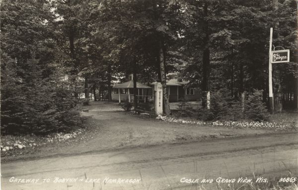 View of Gateway to Bobynn on Lake Namakagon, labeled as Cable and Grand View, Wisconsin. In front is a dirt road with two signs that read "The Bobynn." Behind the entrance gate is a single story building with a porch.