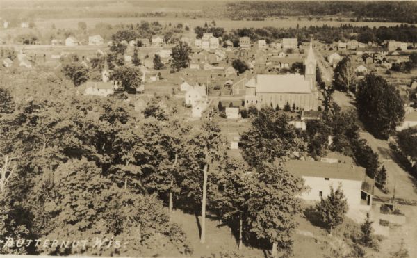Aerial view of town looking down from hill lined with trees. In the foreground railroad tracks cross a road. A church building and numerous dwellings are along the streets.