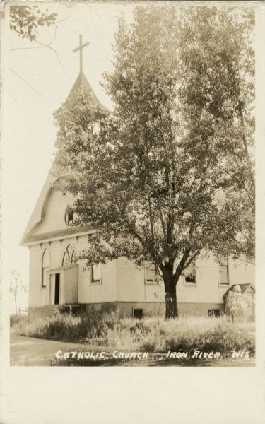 Exterior three-quarter view from road showing the front entrance, with belfry, and a steeple with a large cross at the peak of the roof. There is a large tree and tall grass in the yard surrounding the church.