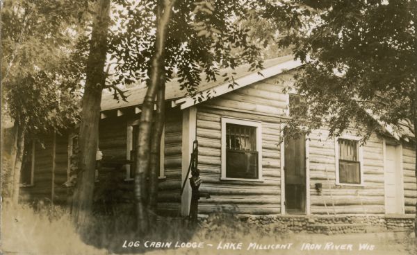 Exterior of Log Cabin Lodge on Lake Millicent, a one-story log cabin with stone foundation with two doors on the right. There is a water pump outside the cabin.