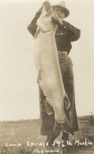 View of Alton Van Camp wearing a hat holding a huge record Muskie listed at "59 1/2 lb" caught by his friend Louie Spray. Caption reads: "Louie Spray's 59½ lb. Muskie, Hayward."