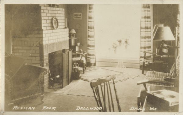 "Mexican Room" at the Bellwood. The room has many chairs, tables and lamps, and a brick fireplace is on the left. Three plants in a holder stand in front of the large picture window framed by striped drapes.
