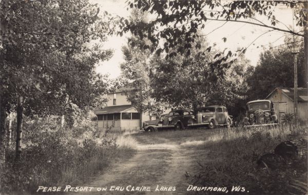 Four automobiles parked at the top of a dirt drive at Pease Resort on Upper Eau Claire Lake. On the far right is a shed or garage, and on the left are trees and a two-story main resort building with a front porch.