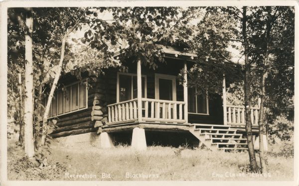 View of a single-story log building with a log front porch labeled as the Recreation Building at Blackburns, Call of the Wild, Eau Claire Lakes.