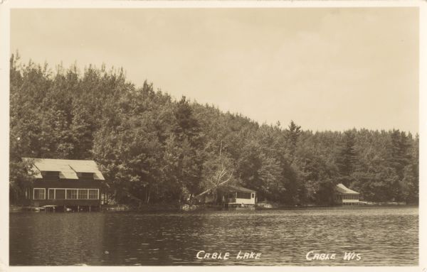 View across lake of three cottages surrounded by trees. Each cottage has a screened porch and a dock on the shore of Cable Lake. There is a group of children and adults near the cottage in the center.